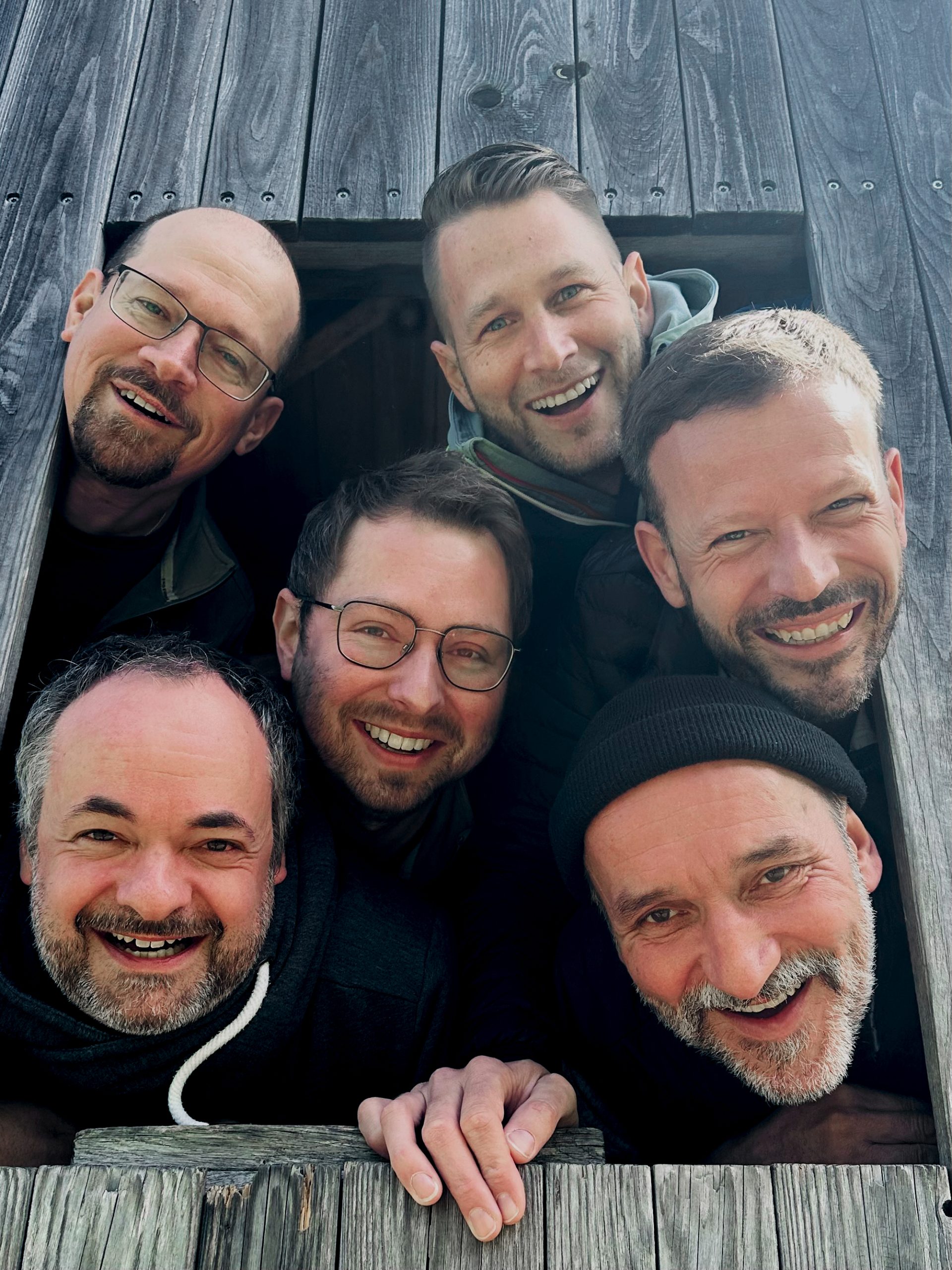 Sechs Männer lächeln fröhlich aus einem Holzfenster, umgeben von rustikalem Holzdesign und natürlichem Licht.