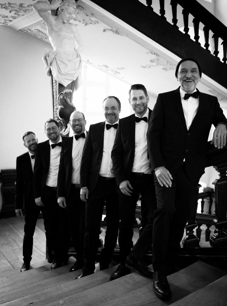 Sechs Männer in Abendkleidung posieren lächelnd auf einer Treppe mit kunstvoller Verkleidung und Skulpturen im Hintergrund.