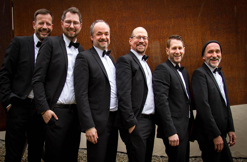 Sechs Männer in eleganten Anzügen mit Fliegen posieren lächelnd vor einer rustikalen Wand im Freien.